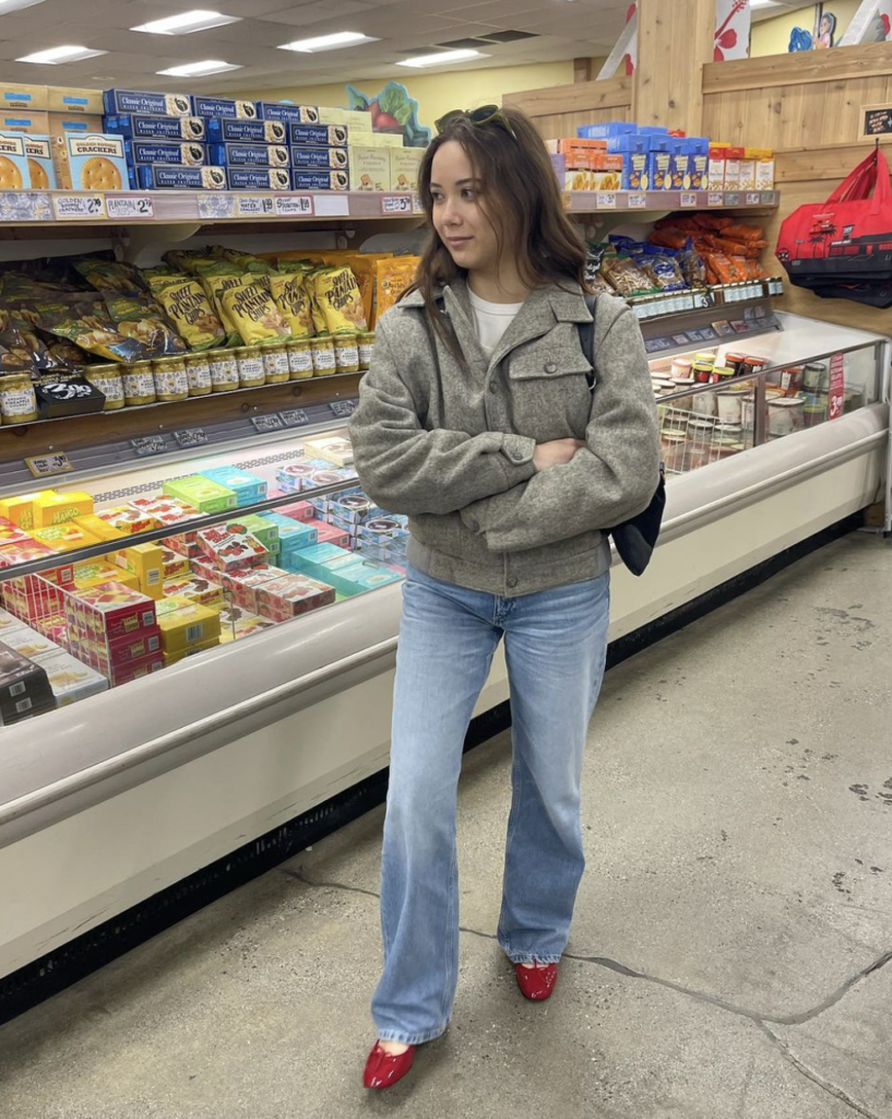 A casual daytime look captured inside a grocery store, with a woman in a grey jacket and blue jeans, accessorized with bold red ballet flats.